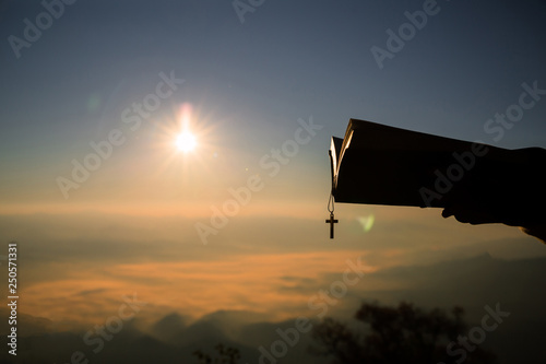 Silhouette of human hand holding bible and cross, the background is the sunrise., Concept for Christian, Christianity, Catholic religion, divine, heavenly, celestial or god.