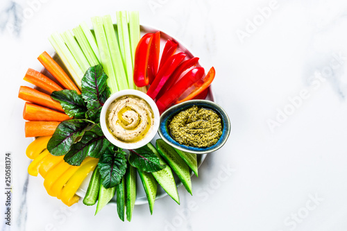 Plate of fresh organic vegetables salad with humus and green pesto dip on white marble background, healthy flat layout food concept with fresh spring or summer vegetables, light dinner