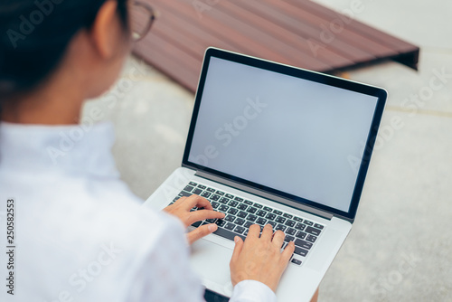 Businesswoman wearing glasses from behind view typing laptop keyboard on her lap with blank space display in front office background- woman office worker and businesswoman concept