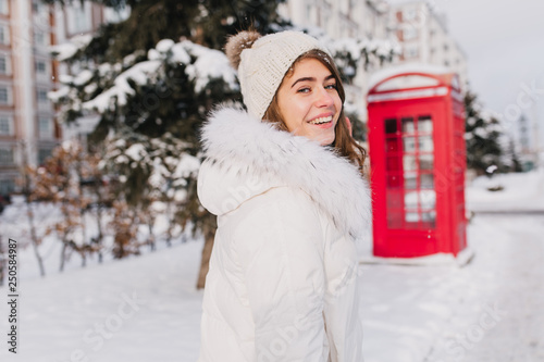 Portrait amazing smiled winter young woman walking on street full with snow in sunny morning. Red telephone box, british style, enjoying cold weather, waiting for christmas