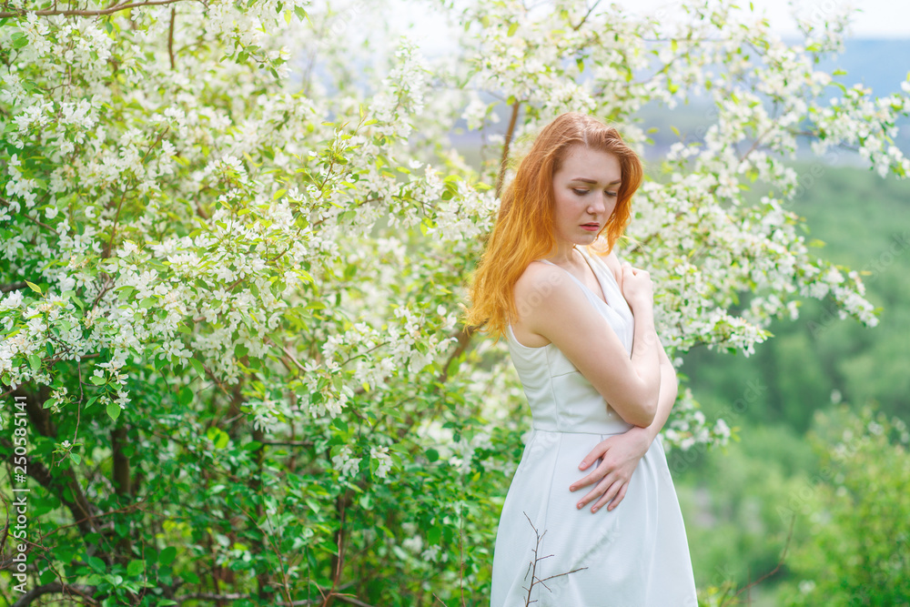 A young and slender girl in the spring stands near a young Apple tree. The sun's rays through the branches of Apple trees, toning. A young and slender girl in the spring stands near a young Apple tree. The sun's rays through the branches of Apple trees, toning.