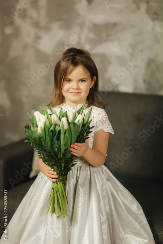 Beautiul little girl hold a bouquet of flowers at home. She sist on sofa at home and waiting for her mom. Bouquet of flowers on 8th of march. Present photo