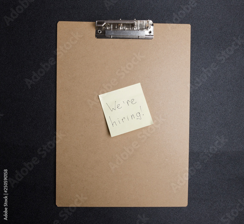 Top view of clipboard and yellow sticker with word hand writing "We`re hiring".Concept of searching for employees