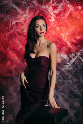 Portrait of magnificent sexy woman in evening dress posing over dark red background