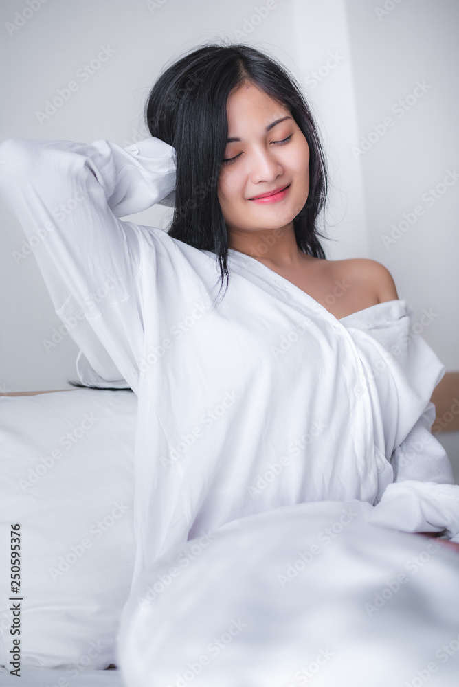 Beautiful young pretty Asian woman wake up and make happy smile with white shirt at the white bed in the morning. Beautiful young pretty Asian woman wake up and make happy smile with white shirt at the white bed in the morning.