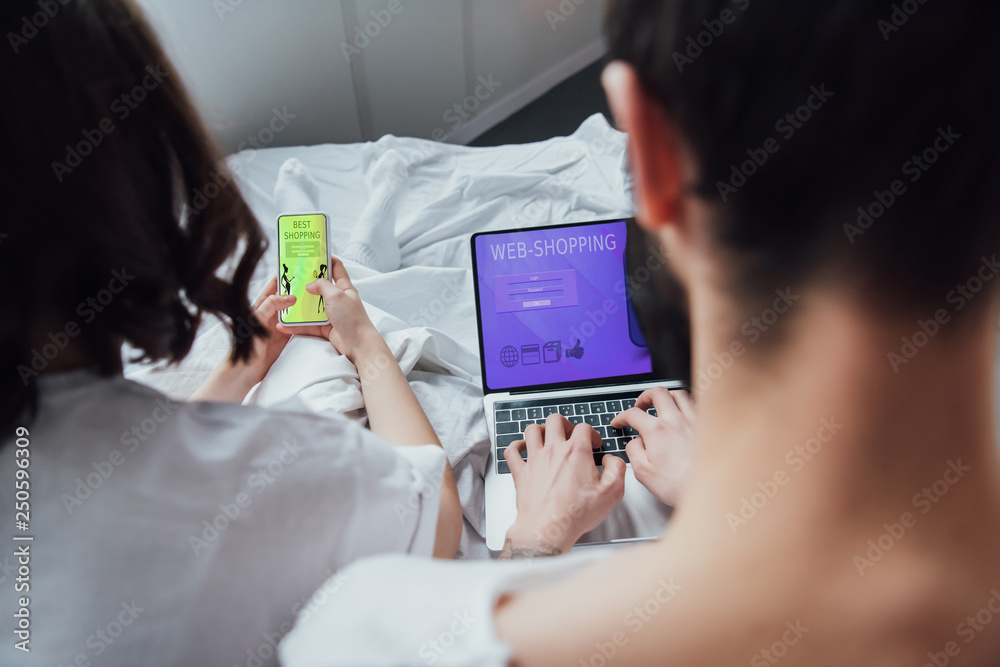 Obraz premium back view of couple lying in bed and using digital devices with online shopping on screen
