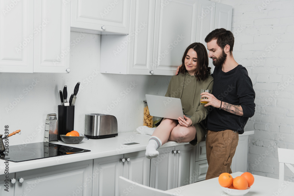 handsome man drinking juice while woman using laptop during breakfast in kitchen handsome man drinking juice while woman using laptop during breakfast in kitchen