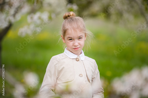 Serious female kid with blonde hair and blowm eyes in the light coat in the blossom gatden. Close-up portrait of 7-8 years old girl. Flowering cherry trees on the background