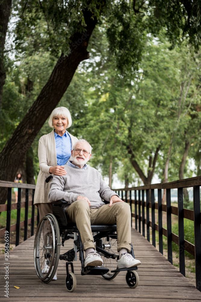 happy senior couple, happy woman with husband in wheelchair on wooden bridge in park
