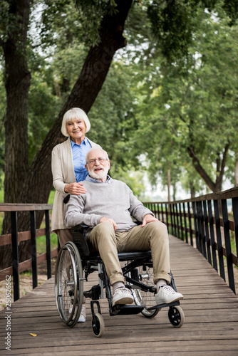 happy woman with smiling husband in wheelchair on wooden bridge in park
