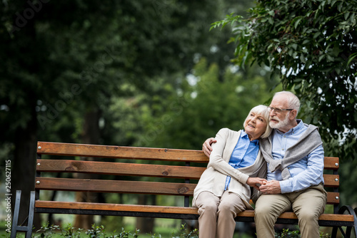 happy senior couple hugging and holding hands while resting on wooden bench in park