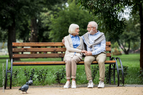 senior couple talking and smiling while sitting on wooden bench in park