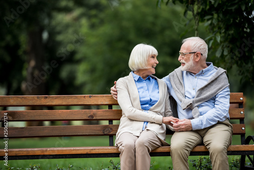 happy senior couple holding hands and looking at each other while sitting on wooden bench park