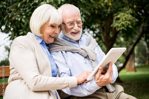 smiling senior couple using digital tablet while sitting on bench in park