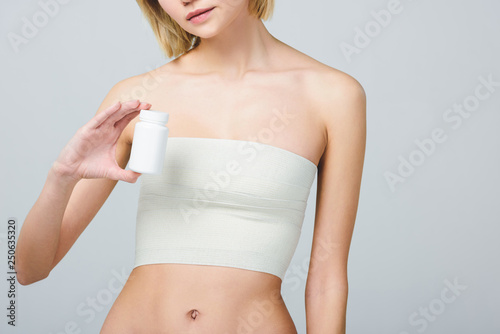 cropped view of woman in bandage on breast after plastic surgery holding pill bottle, isolated on grey