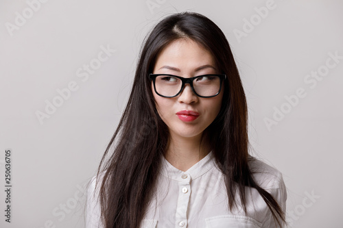 portrait of young calm asian woman in glasses and white shirt looking to the side on gray background. emotions
