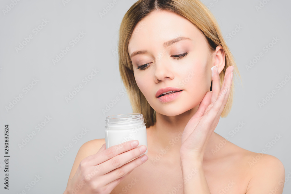 beautiful blonde woman applying cosmetic cream on face and holding plastic container isolated on grey beautiful blonde woman applying cosmetic cream on face and holding plastic container isolated on grey
