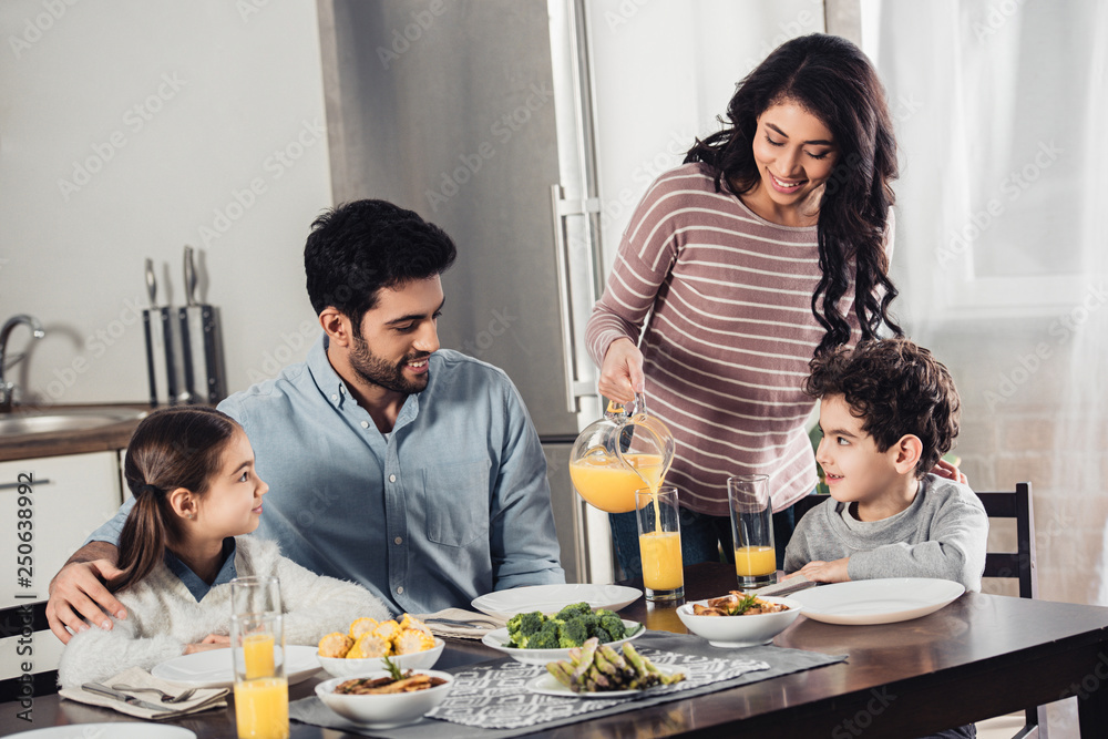 cheerful latin mother pouring orange juice into glass of happy husband near daughter and son during lunch