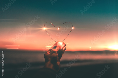 Simbolo del cuore fatto con striscia di luci led tenuto in mano di fronte a un tramonto sull'oceano. San Valentino e festa della donna concetto.
