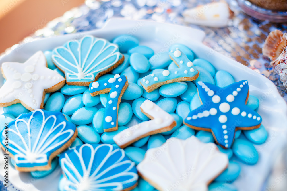 White shell plate full of chocolate candies with blue icing, cookies like shells, stars and number seven and a lot of small jars with marshmallow on the table with shining cloth. Summer season delicio