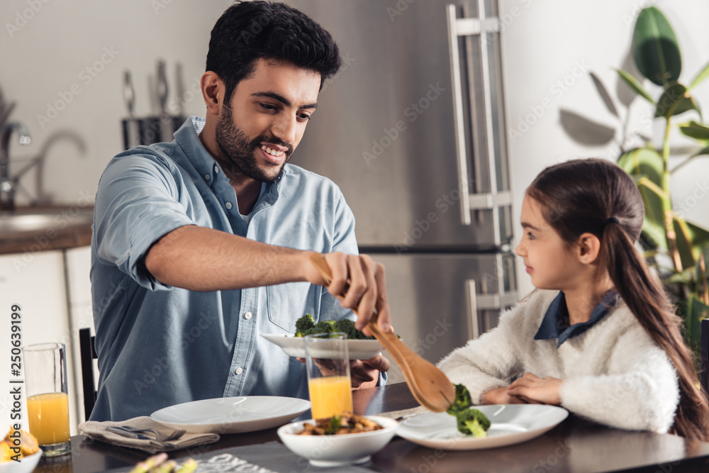 cheerful latin father putting broccoli in plate of cute daughter at home