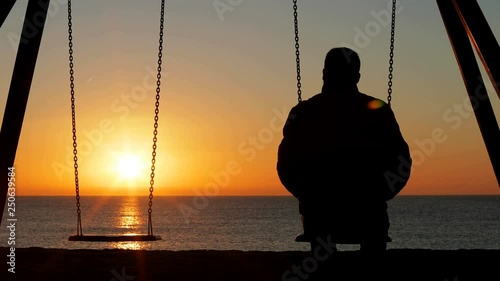 Back view silhouette of a man alone contemplating sunset sitting on a swing on the beach photo