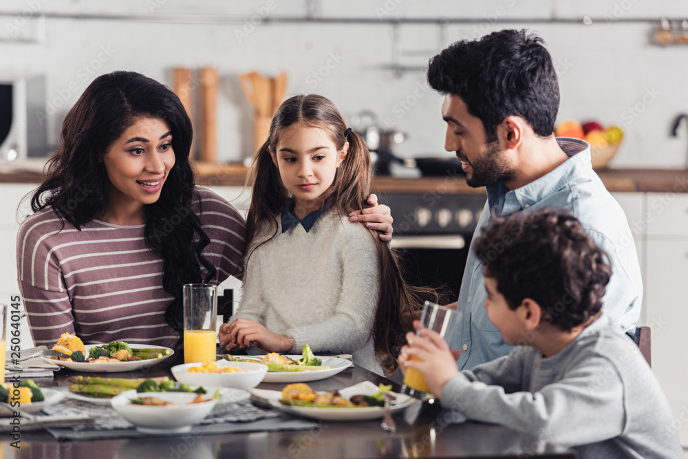 attractive latin mother hugging daughter while looking at son near husband at home attractive latin mother hugging daughter while looking at son near husband at home