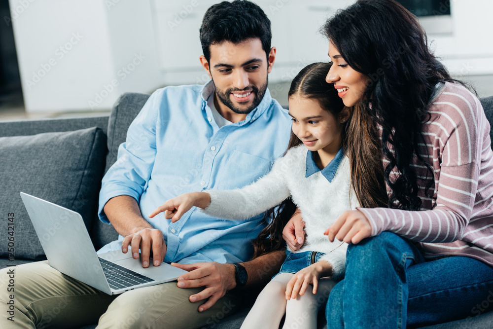 latin woman sitting on sofa with daughter pointing with finger at laptop near dad latin woman sitting on sofa with daughter pointing with finger at laptop near dad
