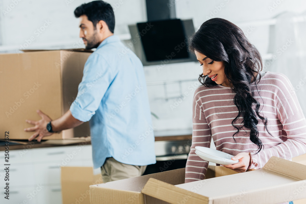 selective focus of attractive latin woman holding plates near boxes with husband on background selective focus of attractive latin woman holding plates near boxes with husband on background