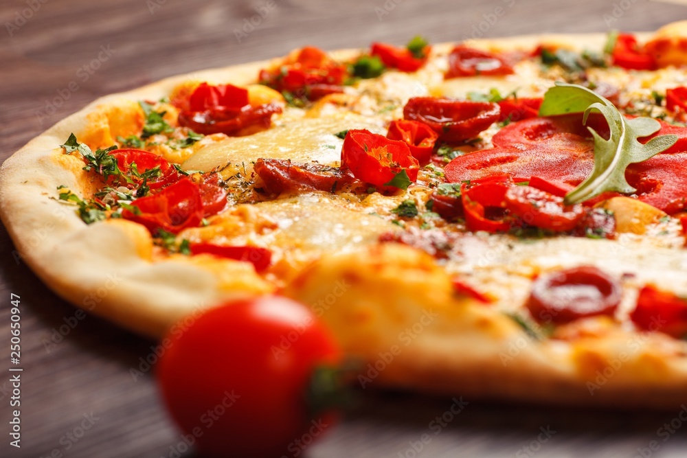 Close up Italian pizza with melted cheese, sausage, snack sticks, pepper and fresh green oregano leaves on a brown table decorated by mushrooms, red sweet pepper and cherry tomatoes