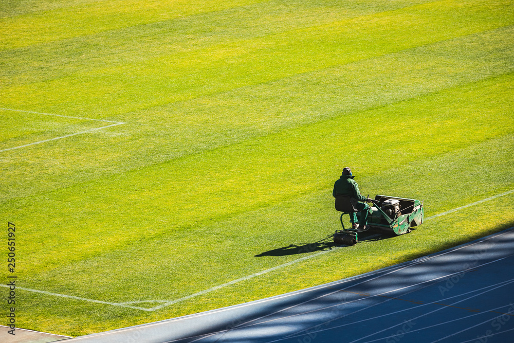 Cutting the grass of the professional football field. Man working with lawn mower in sports stadium amidst the bleachers