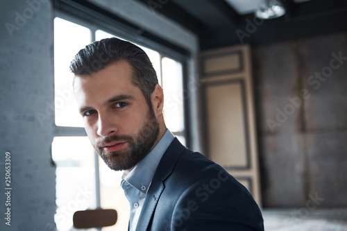 Being successful. Close-up portrait of sexy bearded young businessman who is looking at camera while standing in office