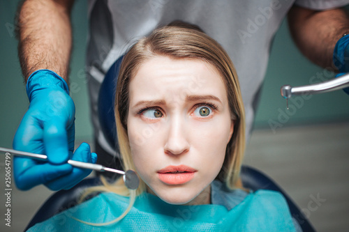 Confused young woman sit in chair in dentistry. She keep eyes rolled. Dentist stand behind her and hold tools for teeth treatment.