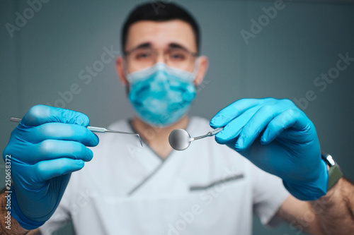 Calm peaceful young make dentist in uniform and mask. He look on camera and hold dental tools. Guy wear latex gloves. Isolated on green background.
