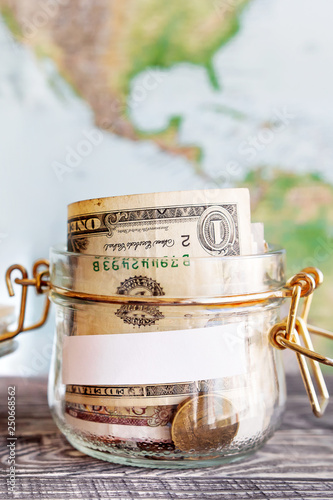 Collecting money for travel. Glass tin as moneybox with cash savings (banknotes and coins) on wooden table and map as background. White empty paper for text.