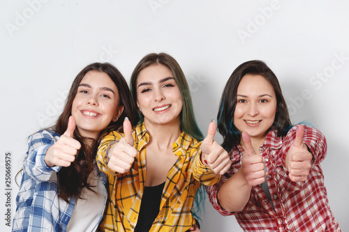Portrait of three beautiful young happy females smiling joyfully showing thumbs up on white