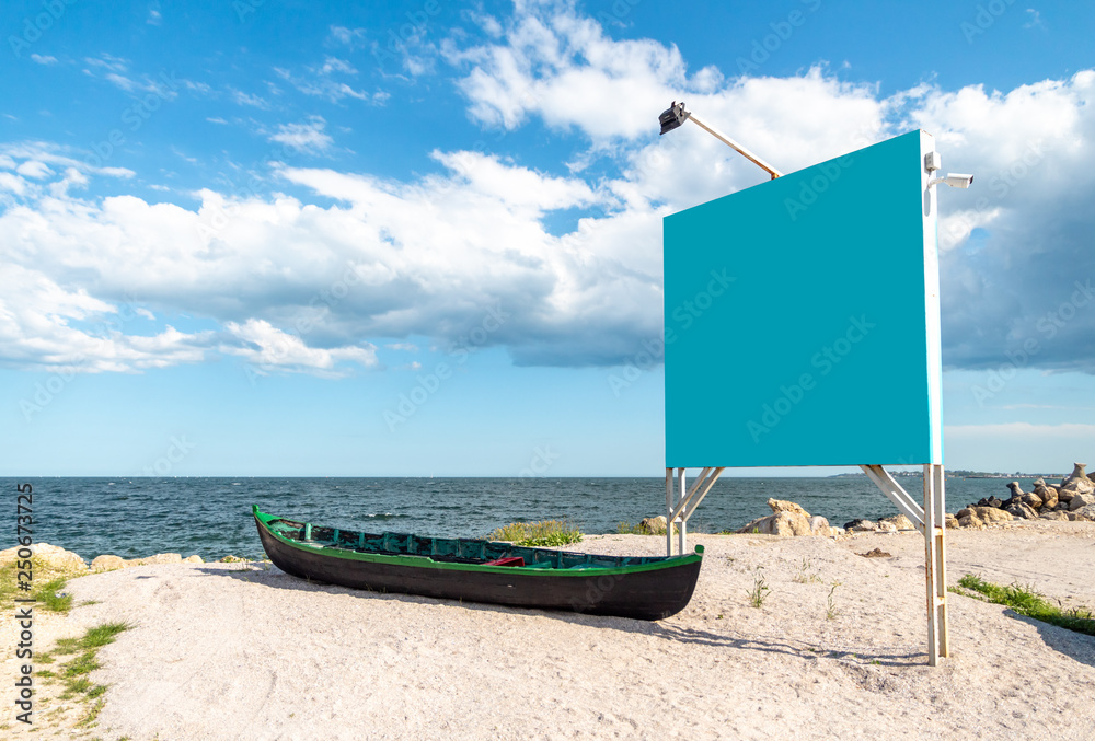 Blue empty billboard next to a local, wooden fishing boat, on the sand, at shore of the Black Sea (Marea Neagra), Romanian coastline. Clear blue Summer sky at the beach.
