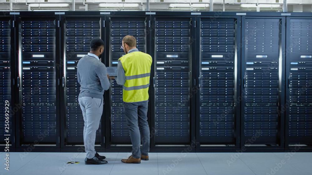 Bearded IT Specialist in Glasses and High Visibility Vest with a Laptop Computer and Black Technician Colleague Talking in Data Center while Standing Next to Server Racks. Running Maintenance. 