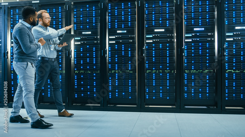 IT Technician with a Laptop Computer and Black Male Engineer Colleague are Talking in Data Center while Walking Next to Server Racks. Running Diagnostics or Doing Maintenance Work.  photo