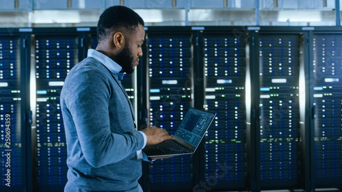 Fotografie Black Data Center IT Technician Walking Through Server Rack Corridor with a Laptop Computer