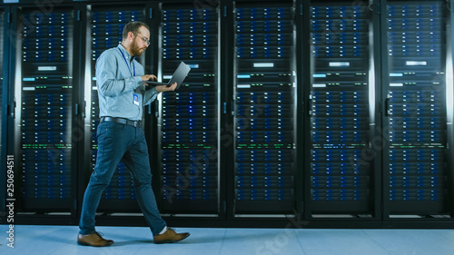 Bearded Data Center IT Professional Walking Through Server Rack Corridor with a Laptop Computer. He Stops and Wirelessly Inspects Working Server Cabinets.