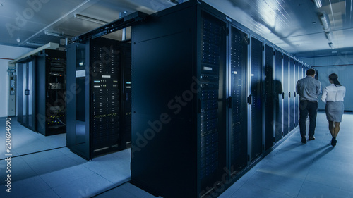 Shot of an IT Admin with a Laptop Computer and Young Technician Colleague Walking Next to Server Racks in Data Center. Running Diagnostics or Maintenance. 