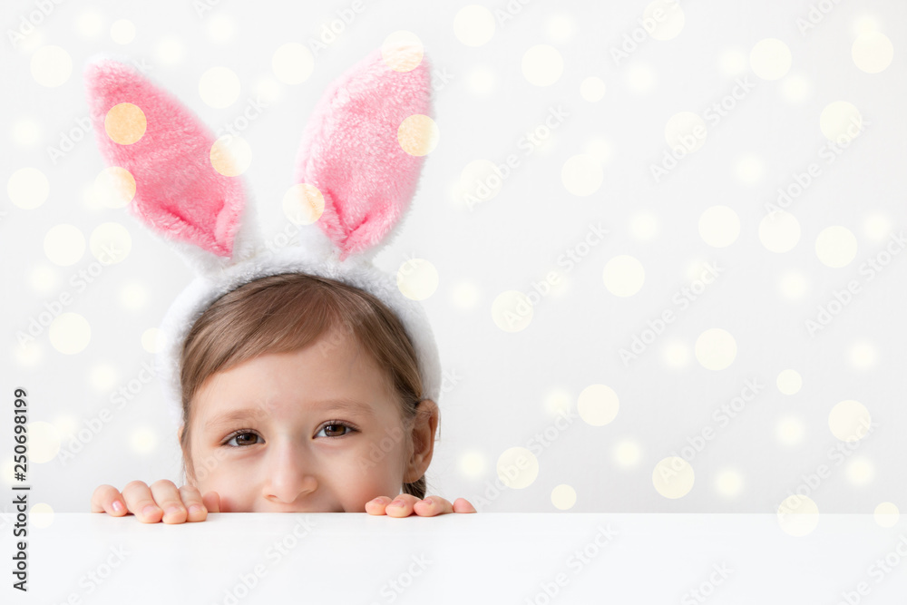 A beautiful little girl with pink bunny ears (hare) is preparing for the celebration of Easter, painting a wooden egg in pink colore. Light background. Free space for text.