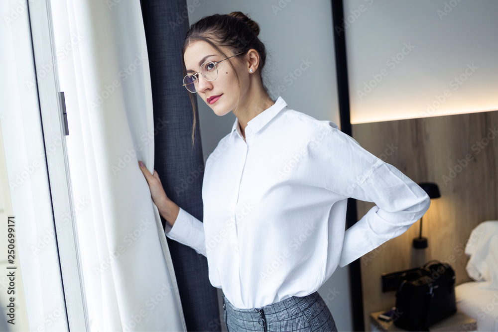 Confident young businesswoman looking sideways standing near the window at hotel room.