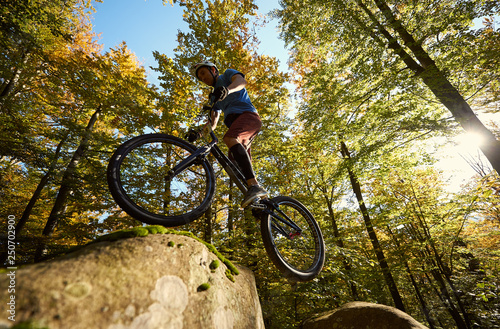 Professional cyclist jumping on trial bicycle on big boulder. Low angle view of male rider making acrobatic trick in the forest on summer sunny day. Concept of extreme sport active lifestyle