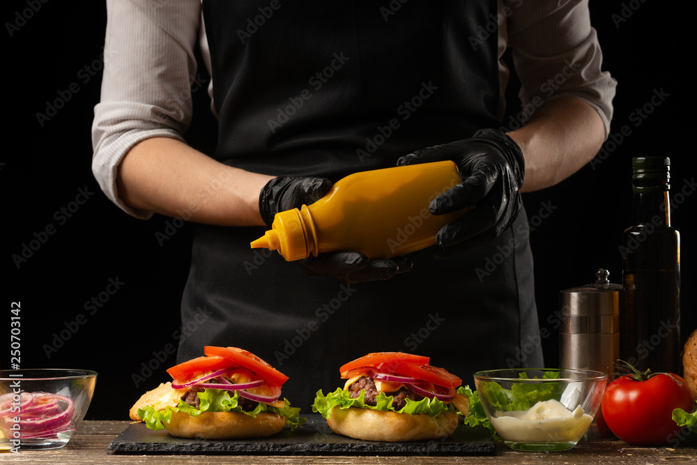 Chef pouring mustard on the burger, on the background of the ingredients. Horizontal photo, Tasty and unhealthy food, fast food, homemade recipes, restaurant, catering, recipe book