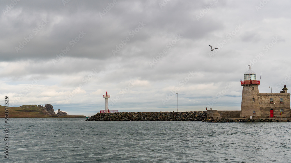 Scene from the beautiful Howth fishing village with a seagull flying over the lighthouse building and the Ireland's Eye Island in the background under a dramatic sky. Seascape from amazing Ireland.