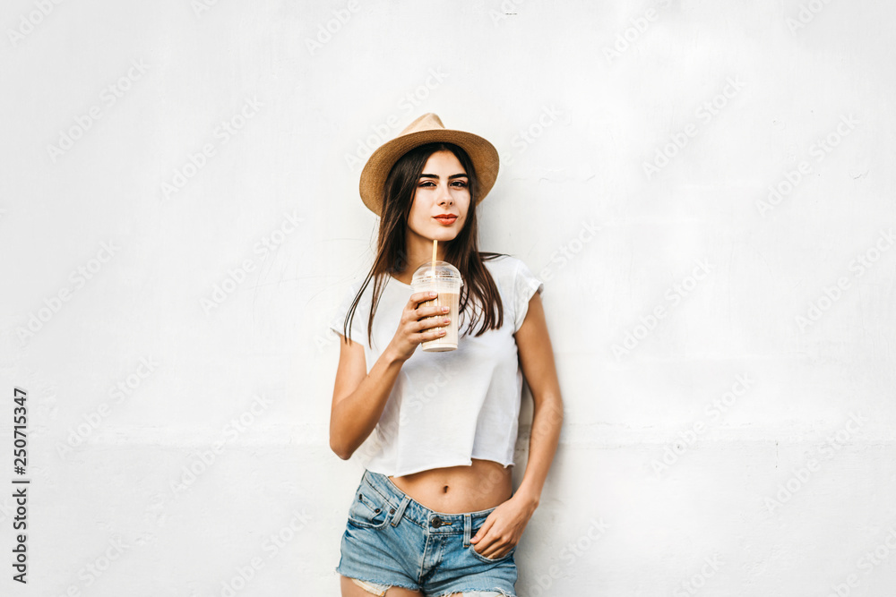 Pretty brunette girl in hat with coffee and white wall behind, copy space.