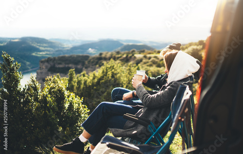 love couple sit in camping chairs on top of sun flare mountain, travelers drink tea on cup enjoy nature, romantic look into distance on background of panoramic landscape, weekend concept photo