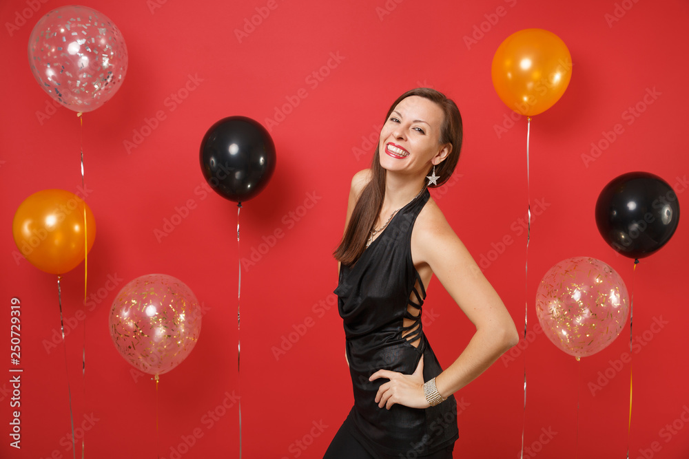 Fototapeta premium Smiling young girl in little black dress celebrating standing with arms akimbo on bright red background air balloons. St. Valentine's, Women's Day Happy New Year birthday mockup holiday party concept.